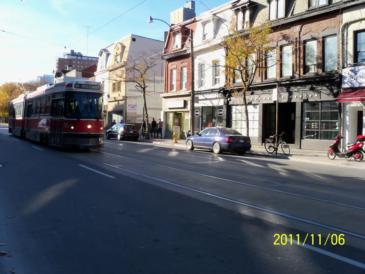 A red TTC streetcar on Queen Street with shopfronts and parked cars along the sidewalk