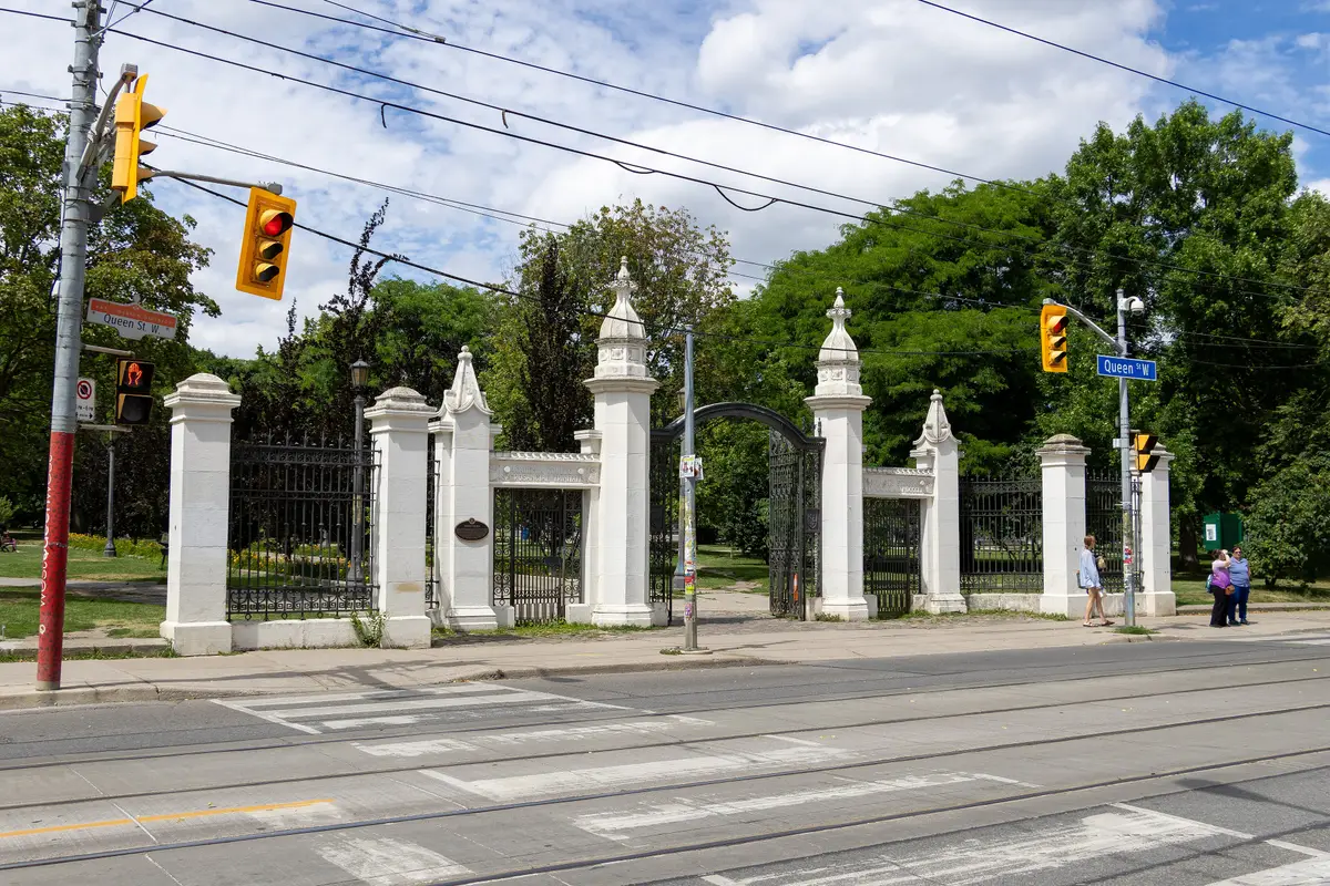 White gateposts and iron railings at the Queen Street entrance to Trinity Bellwoods Park, with a traffic signal on the corner