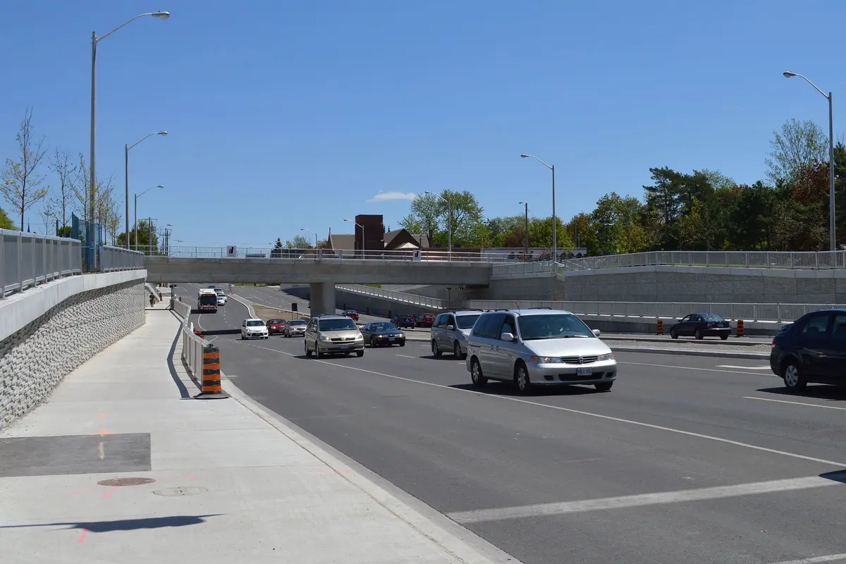 Traffic passing beneath a concrete railway bridge, with a stone retaining wall and sidewalk on the left