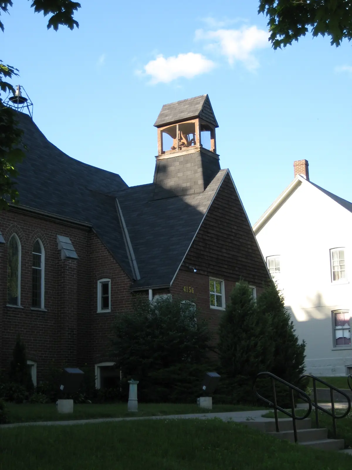 Red-brick church with a dark shingled roof and a small wooden bell cupola on top, beside a white house
