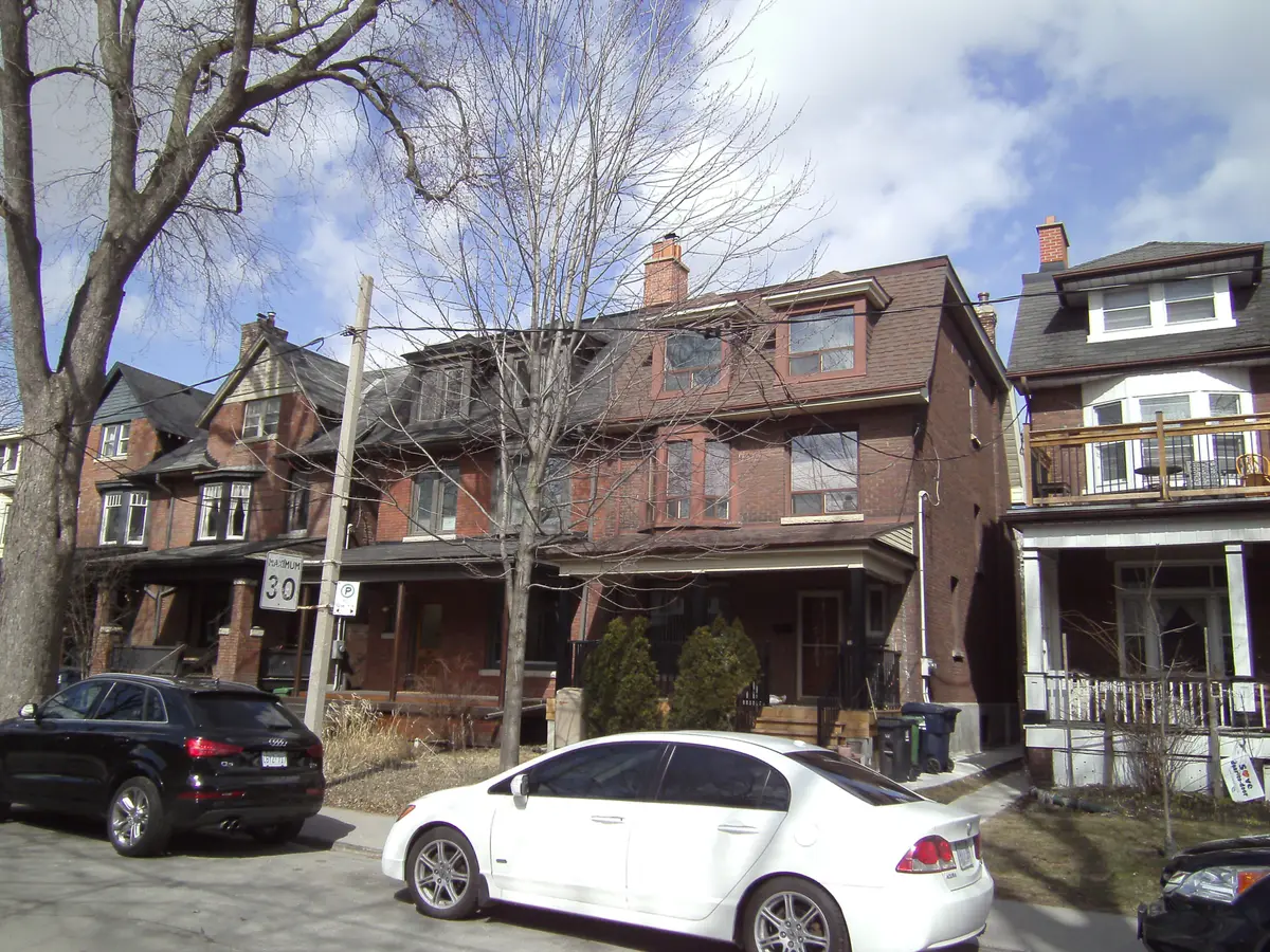 A red-brick house with a mansard roof and a front porch on a tree-lined street, with cars parked out front.