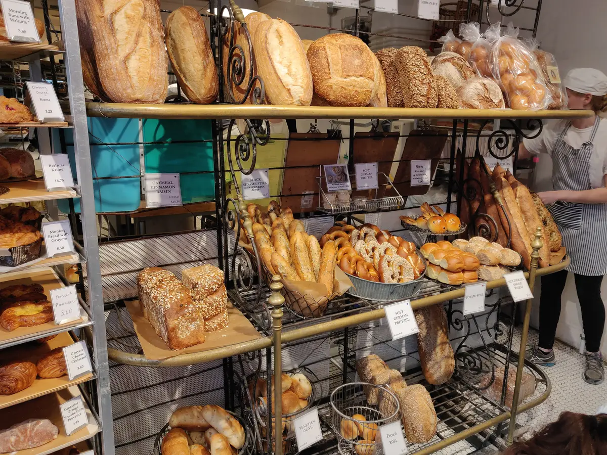 Shelves of loaves, rolls and pastries behind a bakery counter, with a staff member in an apron at the right
