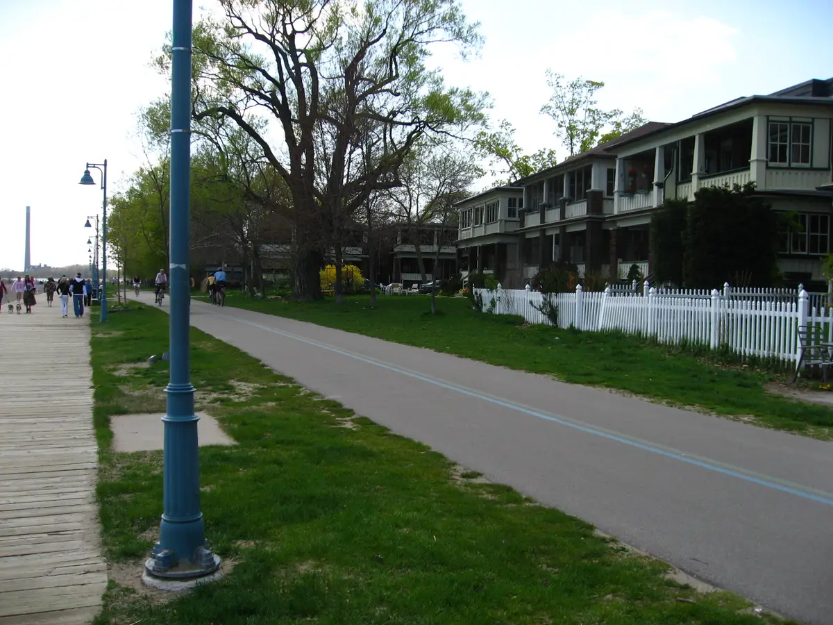 A wooden boardwalk on the left and a paved bike path beside a row of large houses with a white picket fence