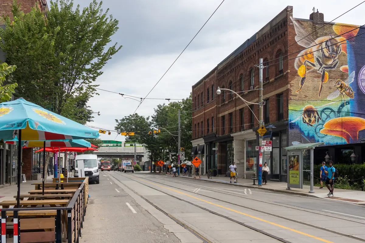 Queen Street East in Riverside, with patio tables and umbrellas on one side, a brick building with a large mural on the other and streetcar tracks between