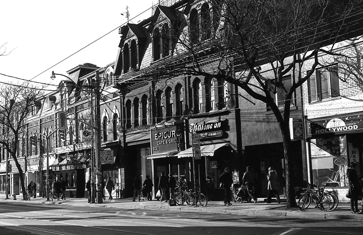 Black and white view of a busy Queen Street West block, with old brick buildings, shopfronts and pedestrians on the sidewalk