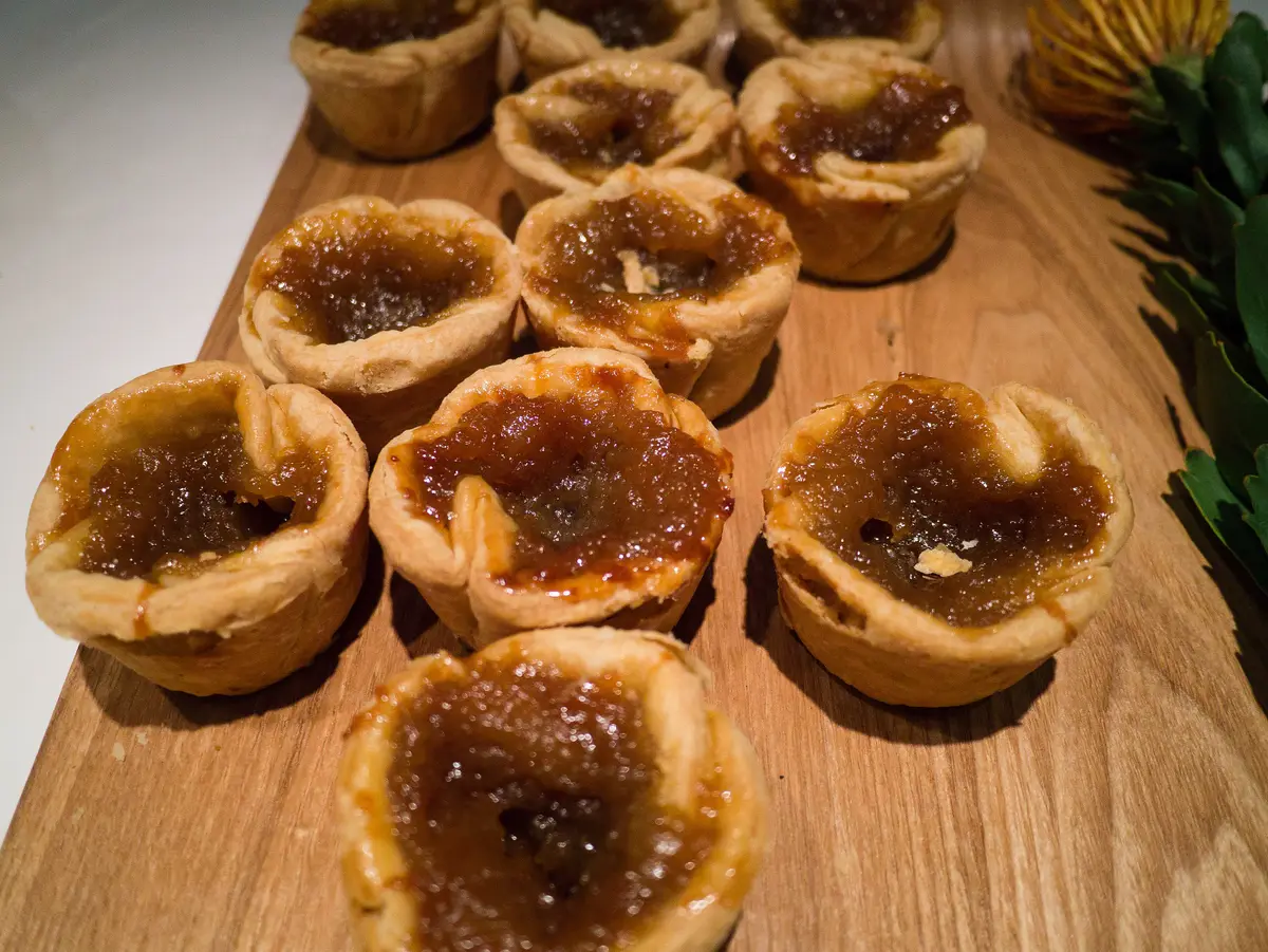 A wooden board of golden butter tarts with crimped pastry edges and glossy filling