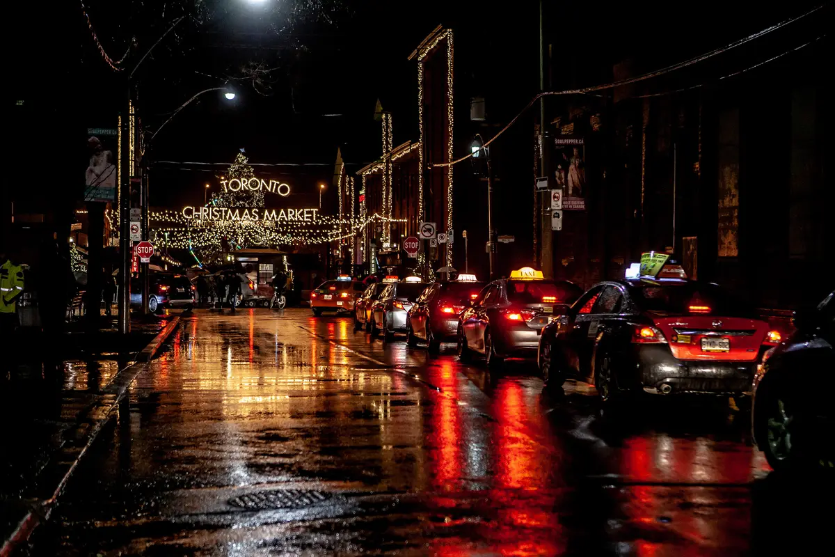 A lit street at night with a glowing holiday market sign, string lights and taxis reflected on wet pavement