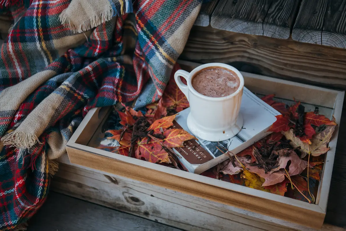 A white mug of hot chocolate on a wooden tray with autumn leaves and a plaid blanket