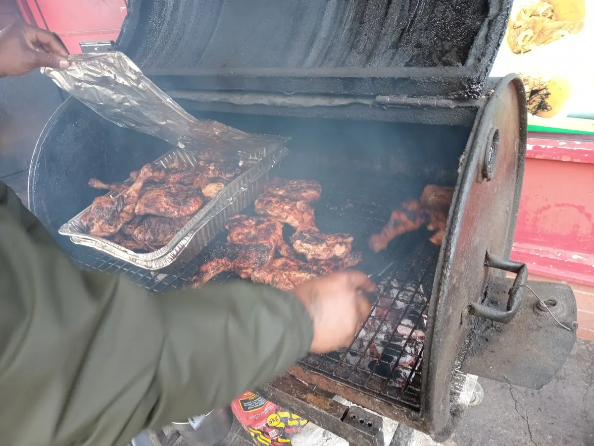 Chicken pieces cooking over coals in a steel drum barbecue while a hand lifts a foil tray of cooked chicken