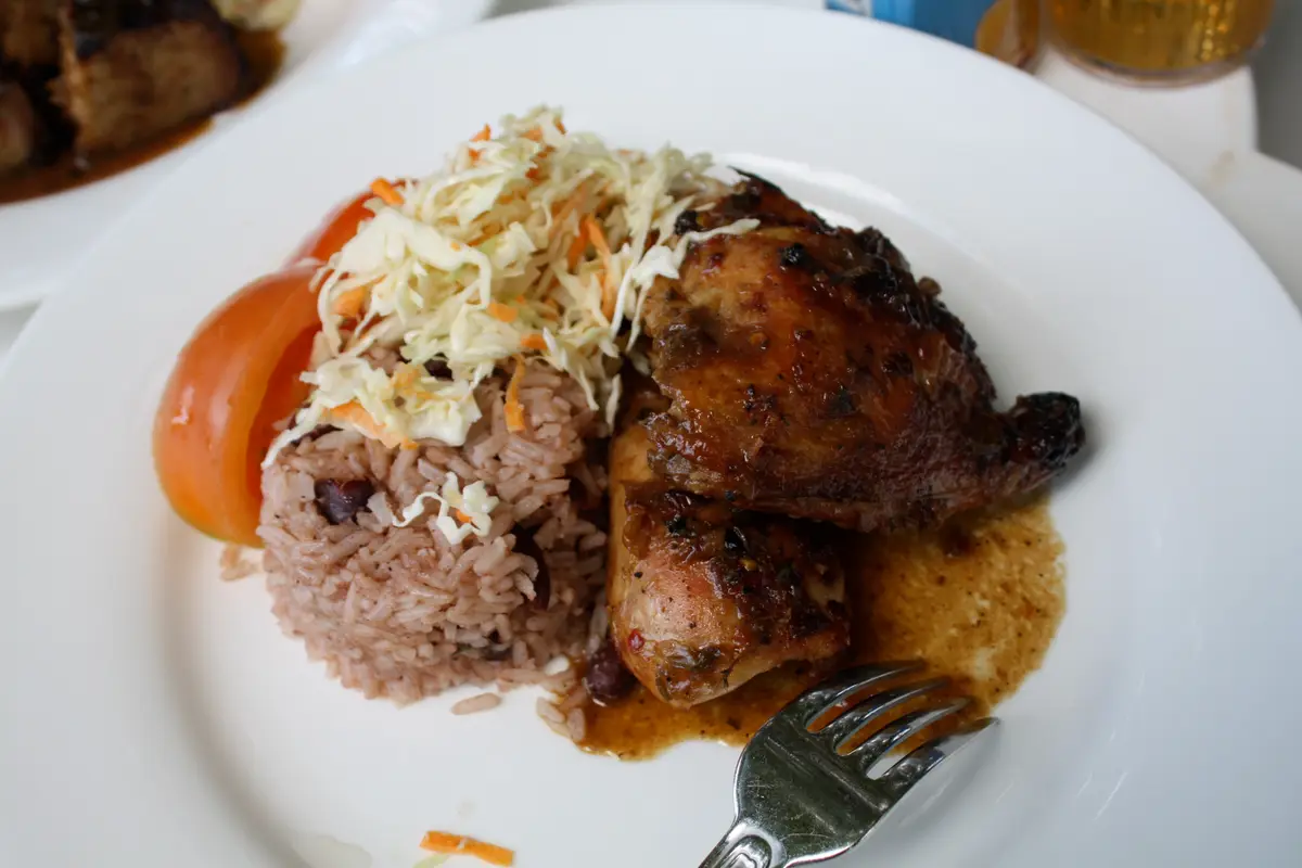A white plate with a jerk chicken leg, rice and beans, slaw and a tomato slice