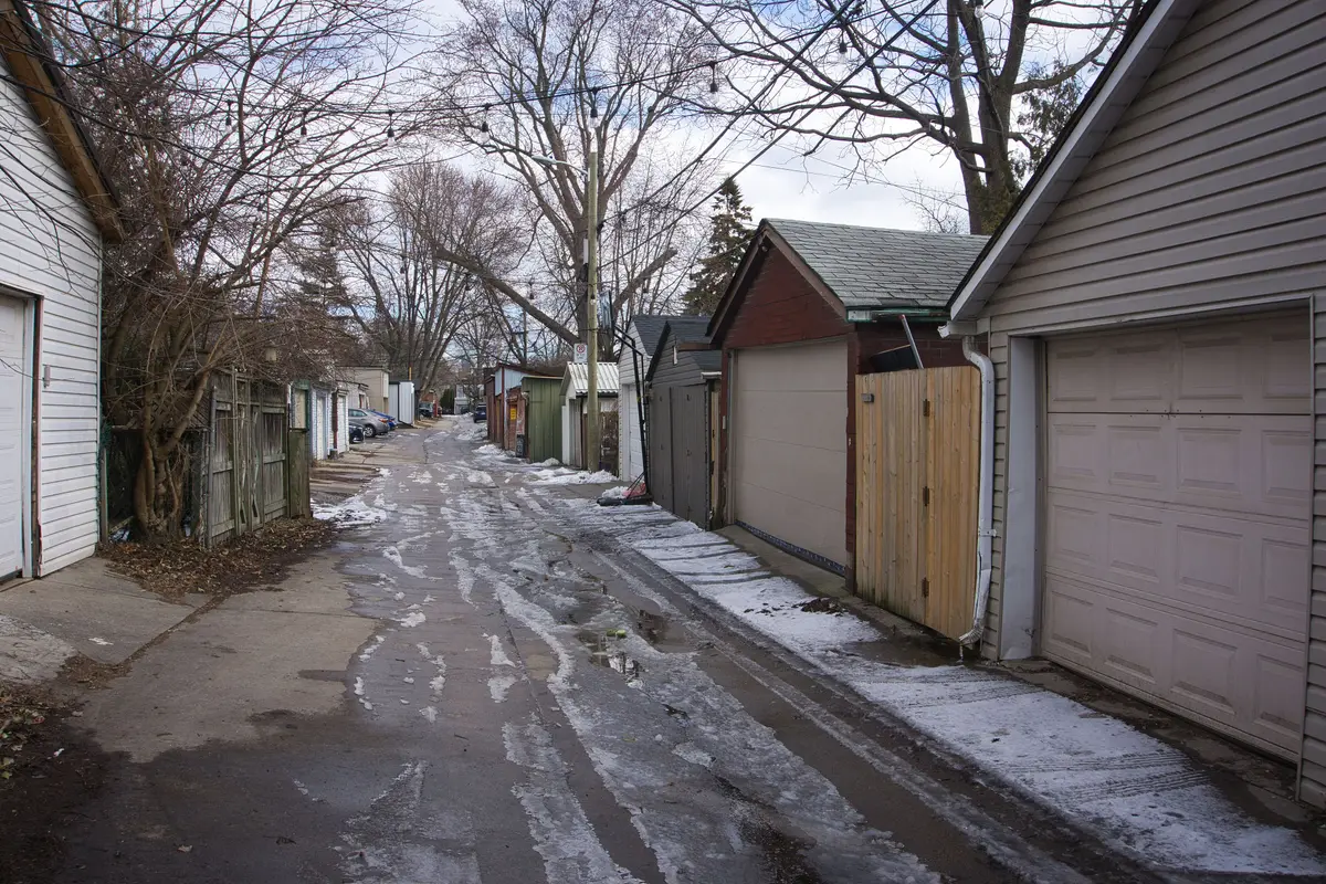 A laneway lined with garages and bare trees, with patches of snow and slush on the ground