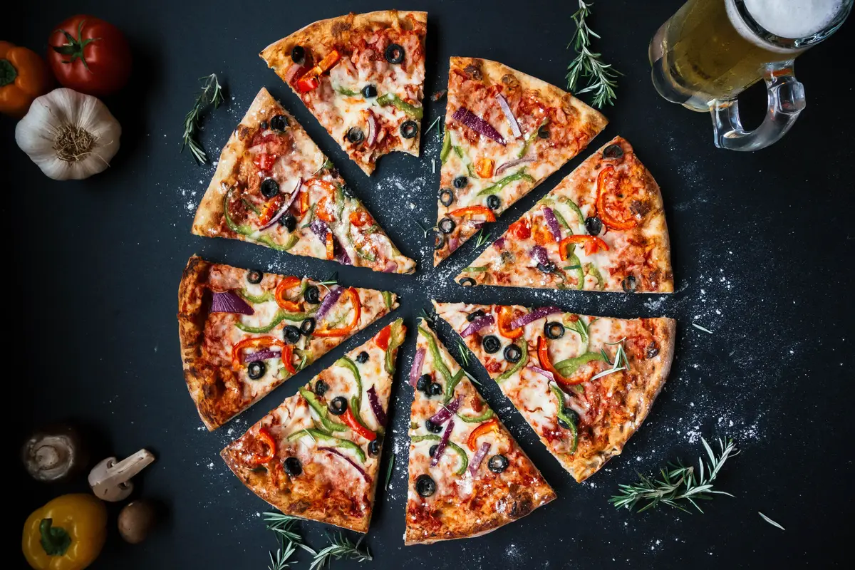 A pizza cut into eight slices on a dark surface, with a glass of beer and fresh herbs beside it