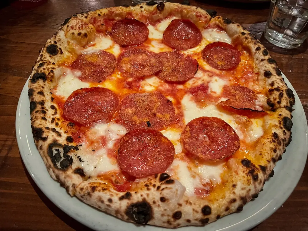 A wood-fired pizza topped with pepperoni and mozzarella, with a puffed, charred crust, on a wooden table