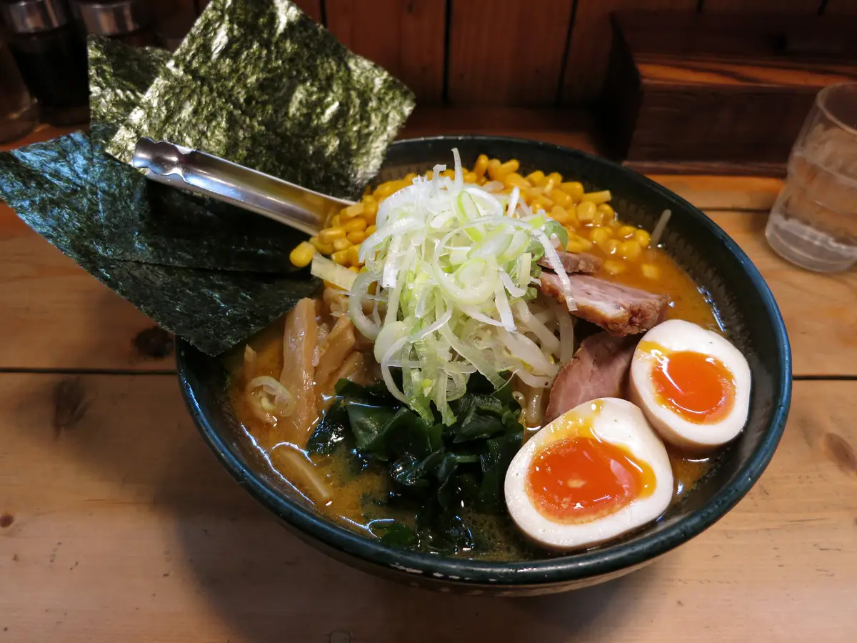 A dark bowl of ramen with halved soft-boiled eggs, corn, green onion, sliced pork and sheets of nori, on a wooden counter.