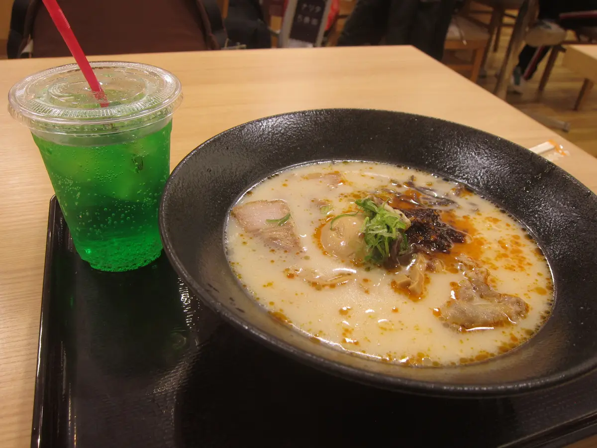 A bowl of creamy tonkotsu ramen with sliced pork and green onion, beside a glass of green soda.