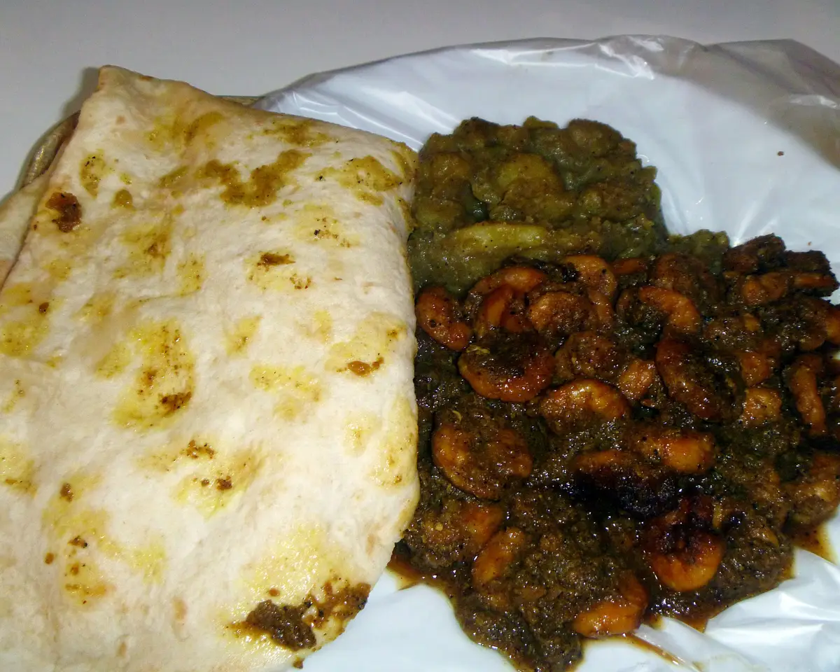 A folded dhalpuri roti beside a plate of curry shrimp with chana and potato
