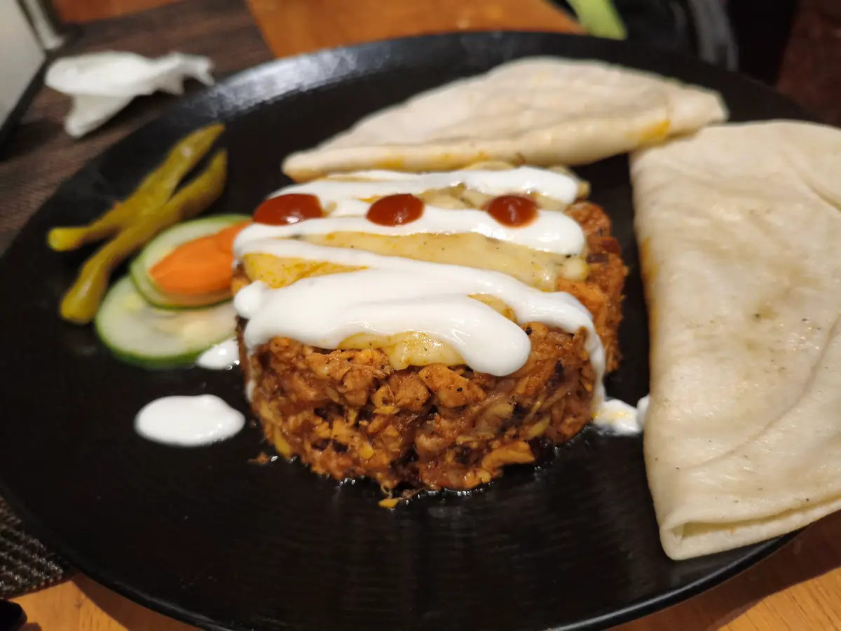 A loaded chicken shawarma plate: shredded meat under white and red sauces, with pickles, cucumber and folded flatbread.