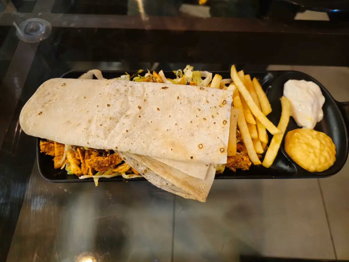 A shawarma plate with a rolled flatbread, shredded meat, fries and creamy dips on a black tray.