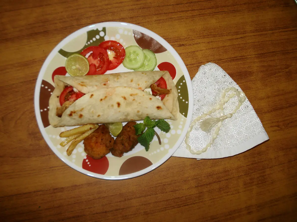 A rolled shawarma wrap on a plate with tomato, cucumber, fries and a lime wedge.