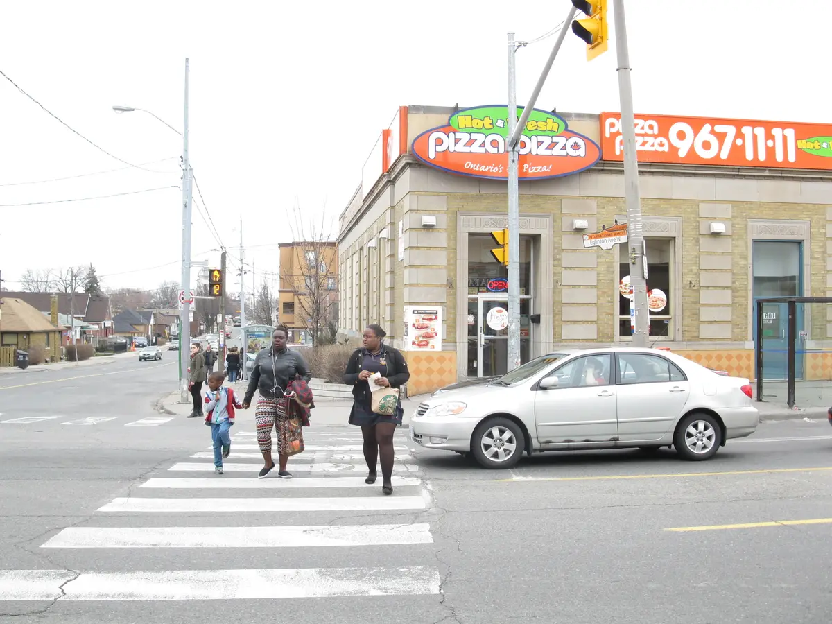 Pedestrians cross Eglinton Avenue at Oakwood Avenue in front of a low commercial building