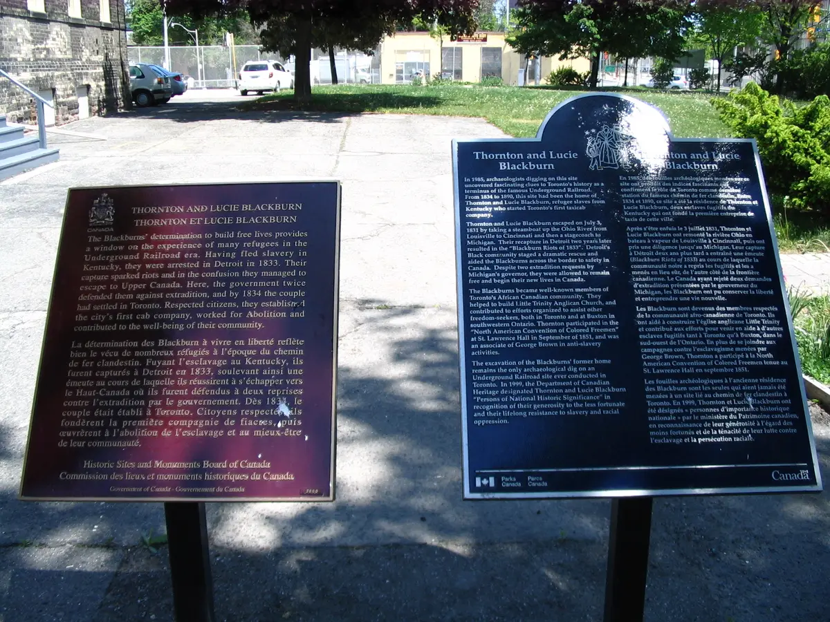 Historic plaques commemorating Thornton and Lucie Blackburn, set on a sidewalk with a lawn behind