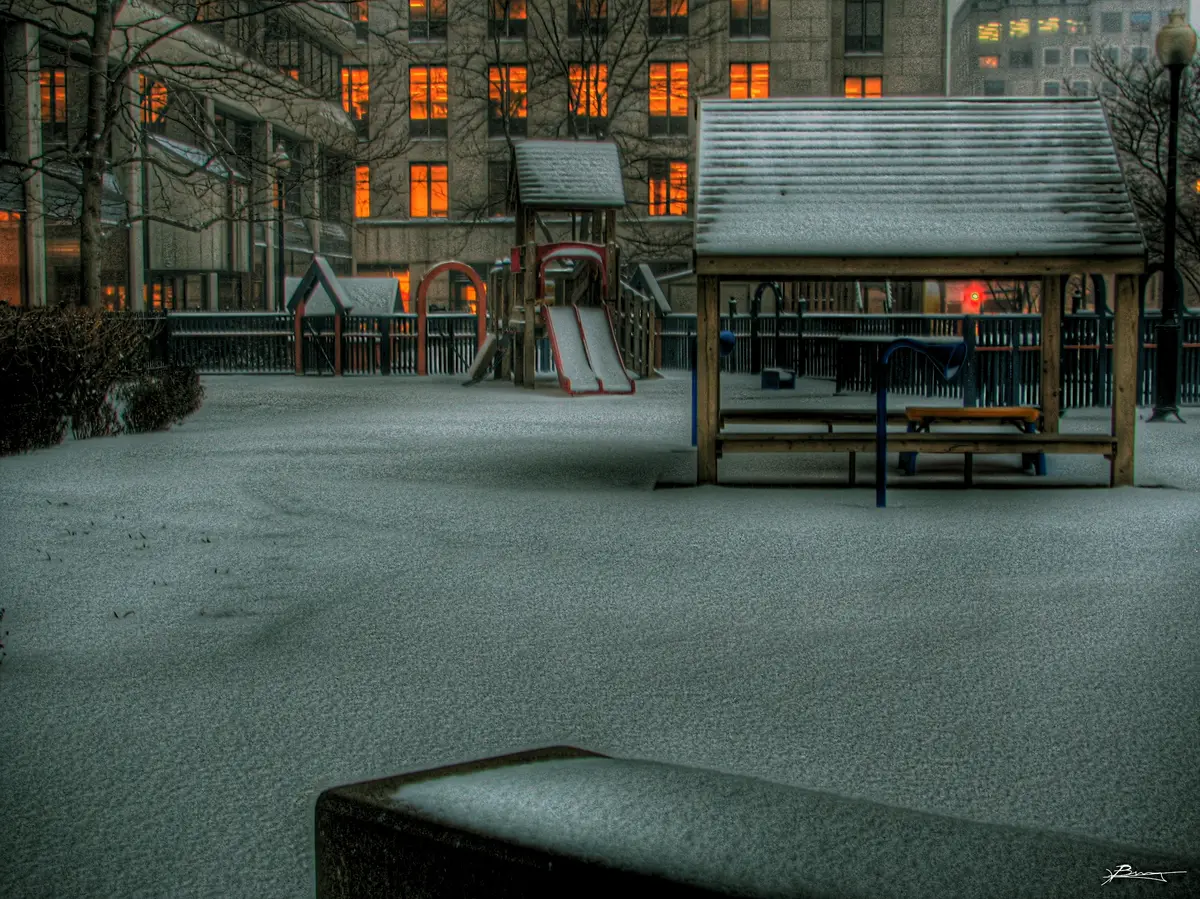 A snow-covered playground at dusk in Toronto, with lit windows glowing in the buildings behind
