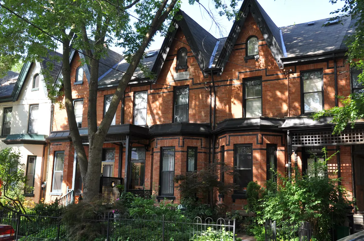 A row of red-brick Victorian houses with steep gables and bay windows under mature trees in Cabbagetown