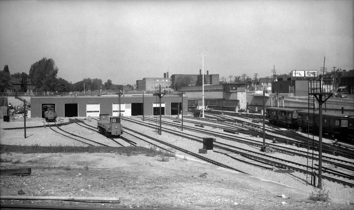 Black-and-white view over a rail yard of many parallel tracks, with a low brick shed with garage doors and two small yard locomotives