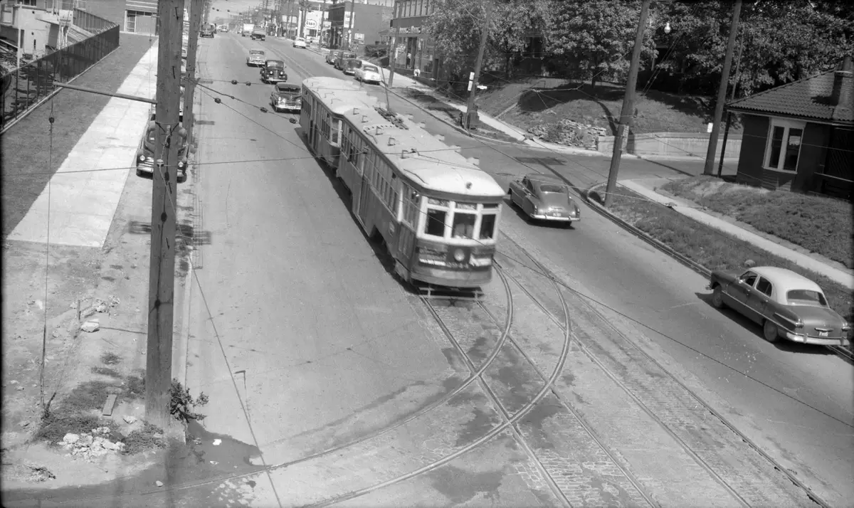Black-and-white view down a wide road from a bridge, with a streetcar curving on its tracks past parked cars and a brick bungalow