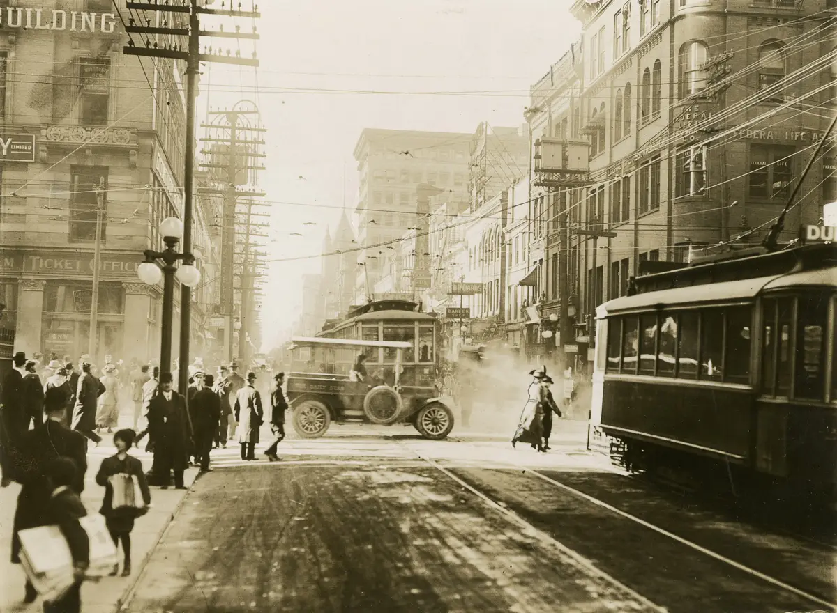 Sepia street scene with a crowd on the sidewalk, an early motor vehicle and a streetcar on a busy downtown street, with brick office buildings and overhead wires
