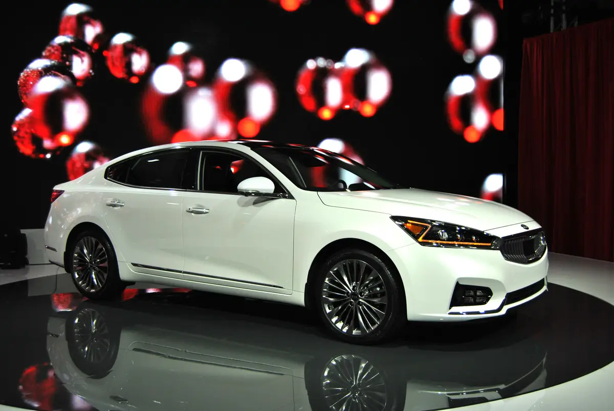 A white Kia Cadenza sedan on a display platform in front of a dark backdrop with red lights.