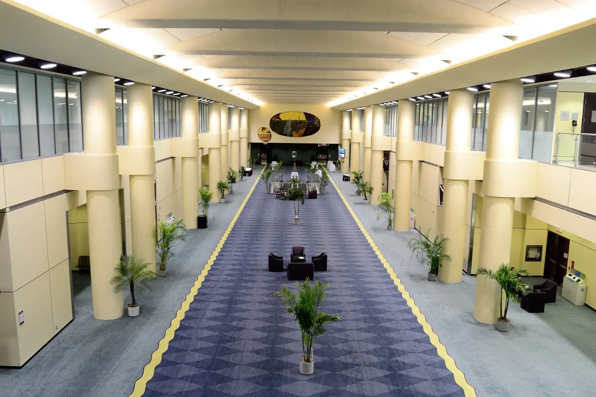 An empty concourse with tall cream pillars, potted palms and a blue patterned carpet.
