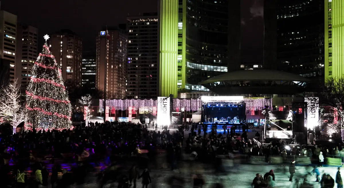 A large lit Christmas tree and a stage with a crowd on the rink at Nathan Phillips Square at night