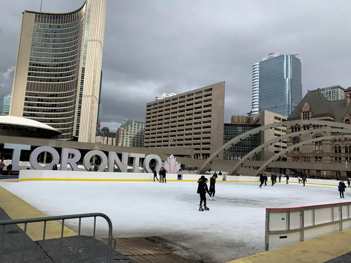 Skaters on the rink at Nathan Phillips Square, with the Toronto sign and City Hall behind them under a grey sky.