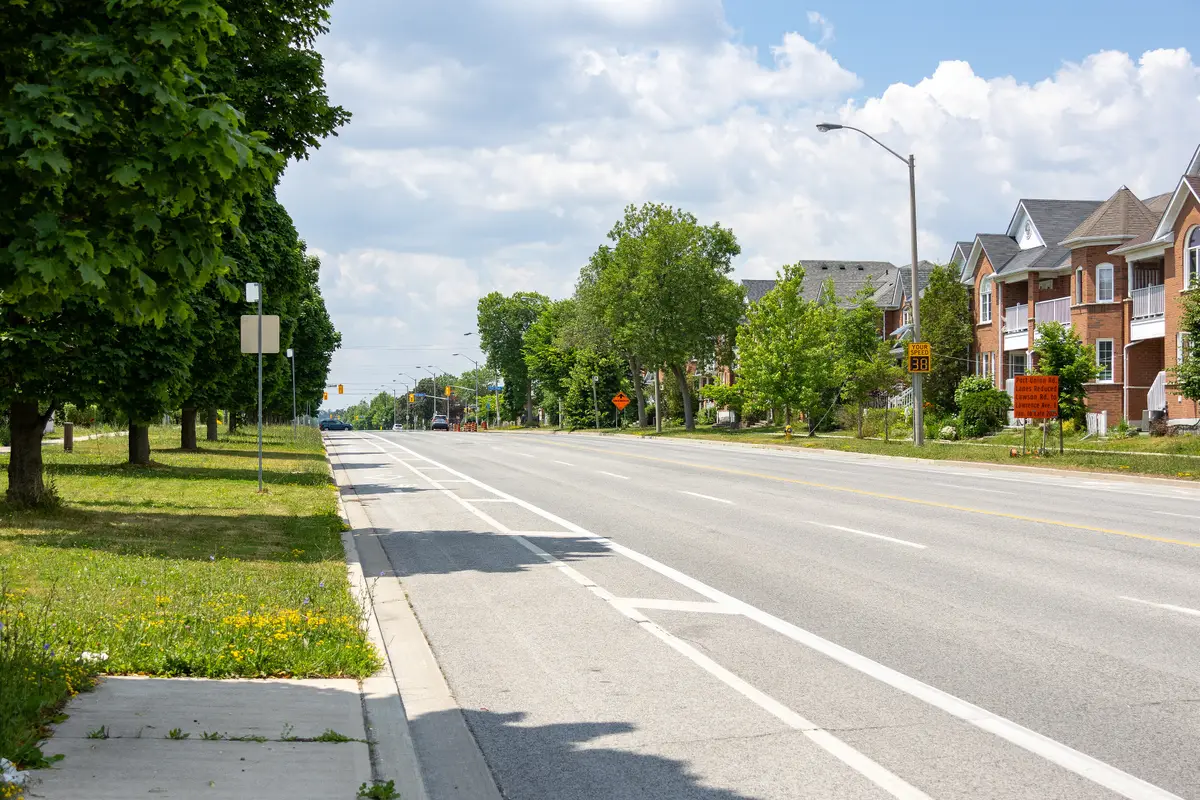 Wide Lawrence Avenue East lined with trees on one side and houses on the other, under a partly cloudy sky