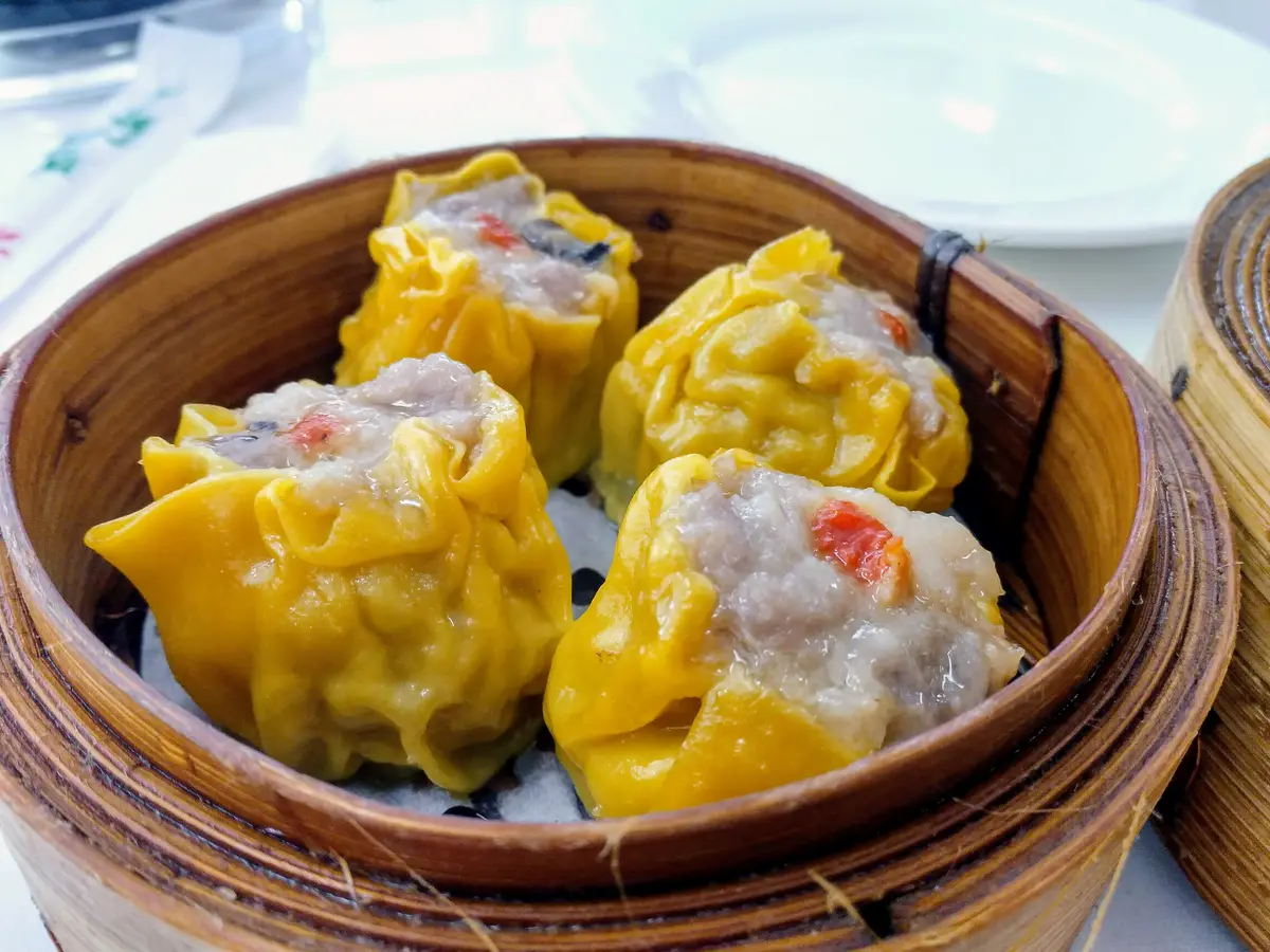 Four dim sum with yellow wrappers, each topped with a small red berry, in a bamboo steamer basket.