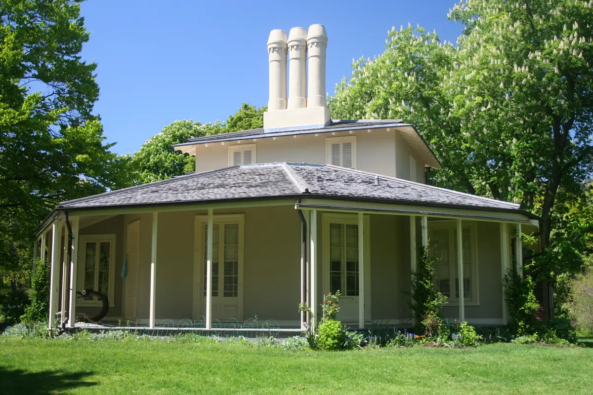 A single-storey Regency-style cottage with a wide veranda and tall white chimneys, set among trees in High Park.