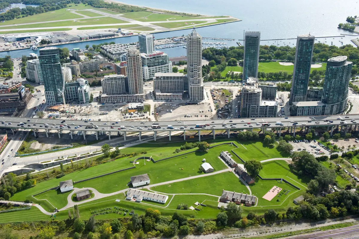 Aerial view of the green Fort York grounds with low heritage buildings, the Gardiner Expressway and condo towers beside the lake.