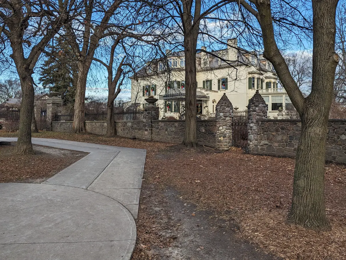A large pale mansion seen through bare trees, behind a stone wall and a paved path.