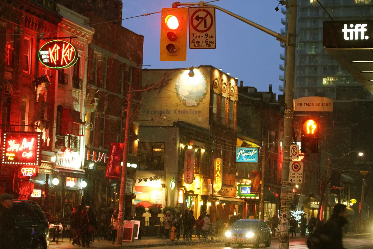 Neon signs and lit shopfronts on a snowy evening street, with a traffic light and a passing car