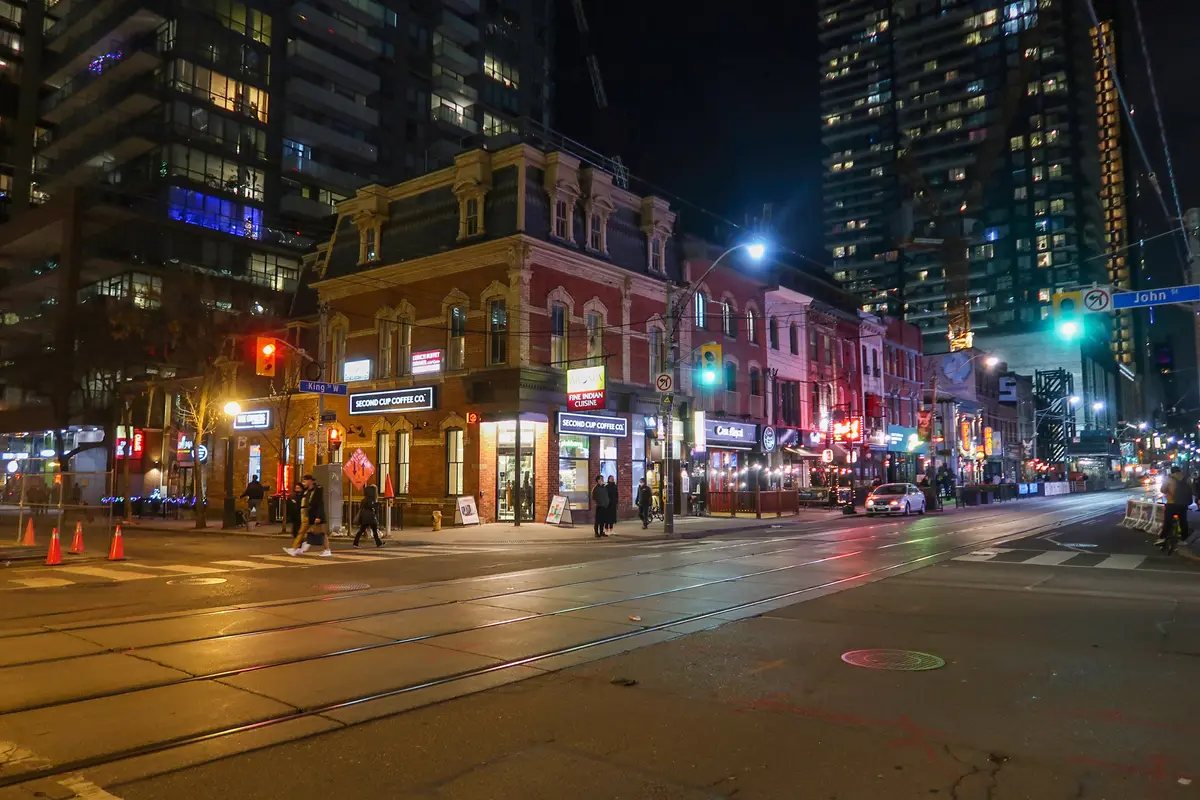 A street corner at night with red-brick buildings, streetcar tracks and lit shopfronts