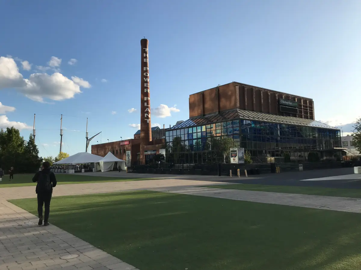 Brick building with a tall chimney marked The Power Plant, beside a glass gallery front and a lawn.
