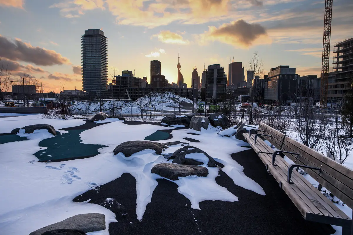 Sunset over snow-covered rocks and a wooden bench at Corktown Common, with towers and the CN Tower beyond
