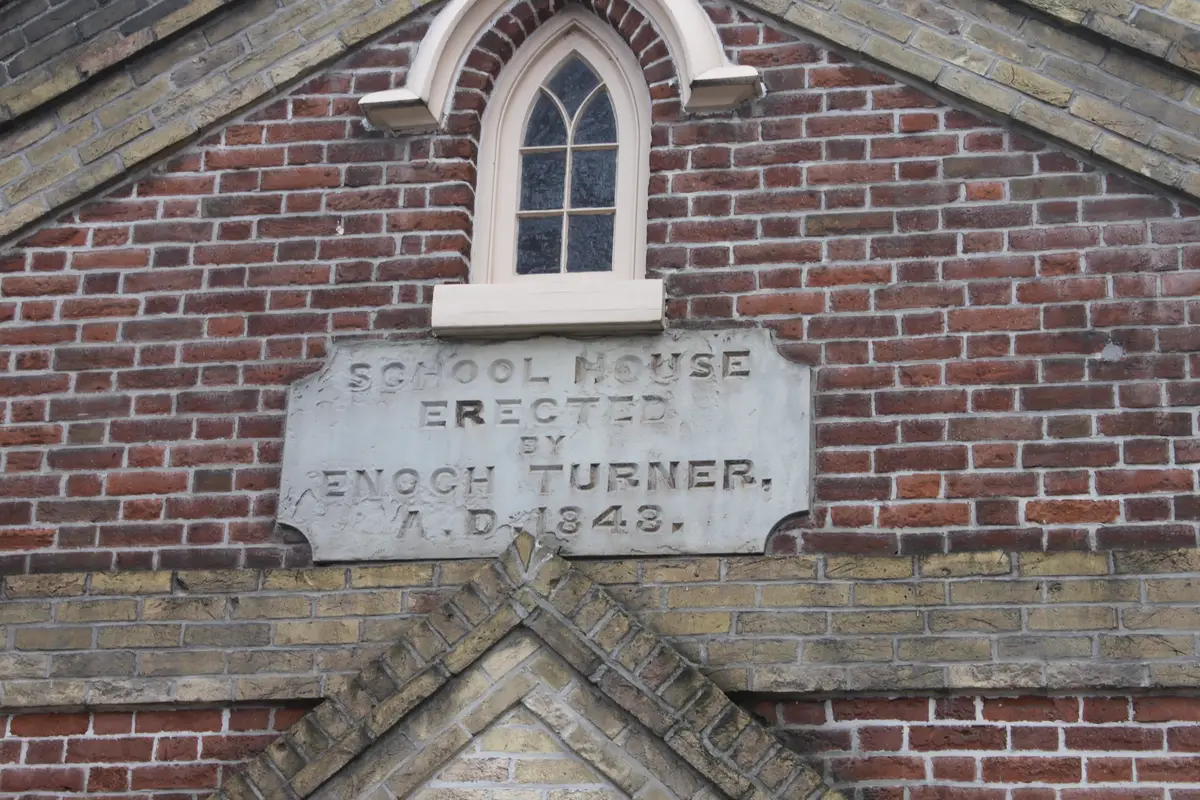 A stone plaque on the brick gable reads School House Erected by Enoch Turner, below a pointed arched window