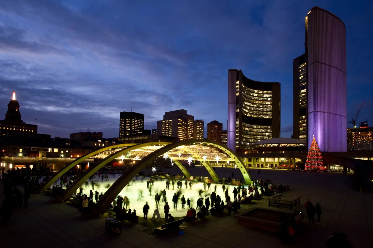 Skaters on the lit rink at Nathan Phillips Square at night, under the arches and beside the City Hall towers