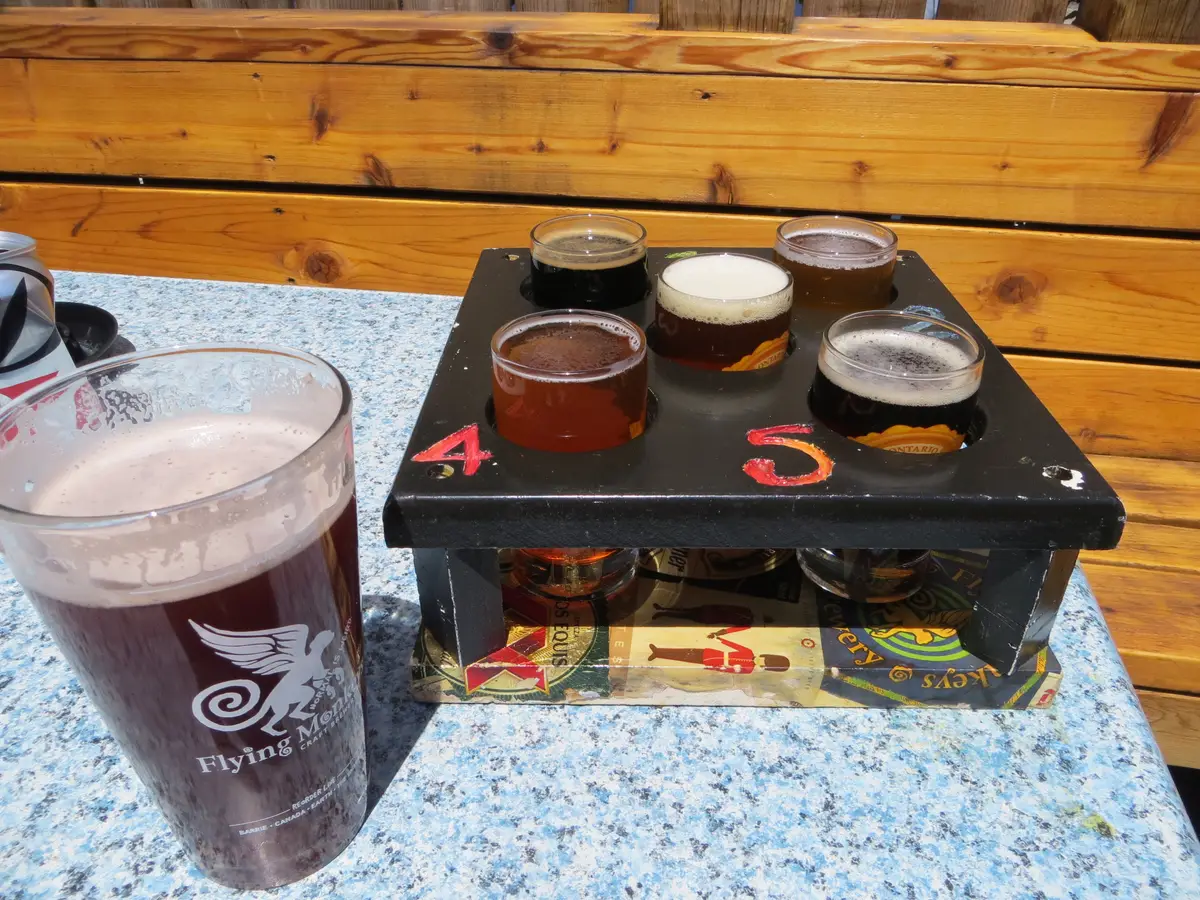 A wooden flight tray of five small beer glasses beside a pint on a sunny patio table