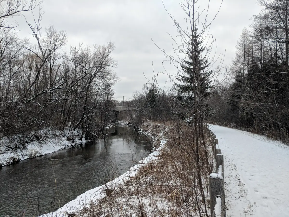 A snow-dusted path beside a creek, with bare trees and a wooden fence under a grey sky