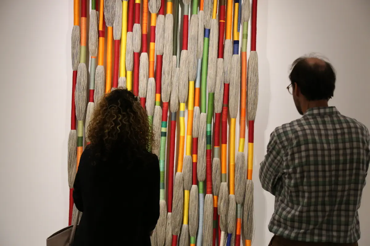 Two visitors seen from behind look at a tall textile hanging of coloured wrapped cords and grey fringed bundles on a white wall