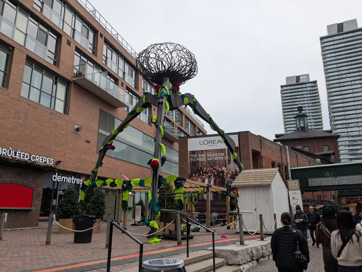 A tall spider sculpture decorated with holly in front of brick buildings and condo towers in the Distillery District
