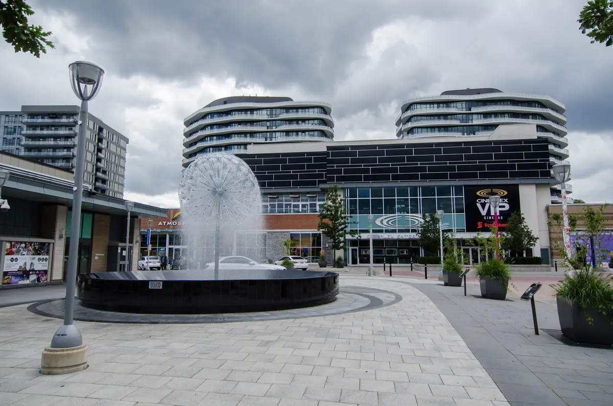 A round fountain on a paved plaza at the Shops at Don Mills, with curved modern buildings and a cinema sign behind
